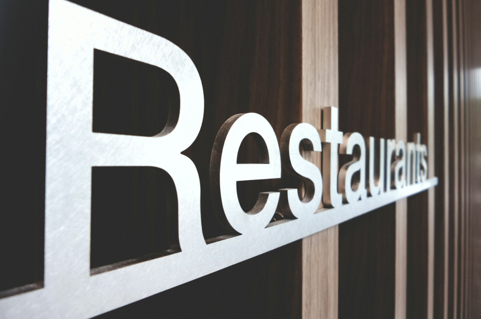 Close-up of a modern "Restaurants" metal sign with a wooden background, angled slightly to the right. The tone is sleek and contemporary.