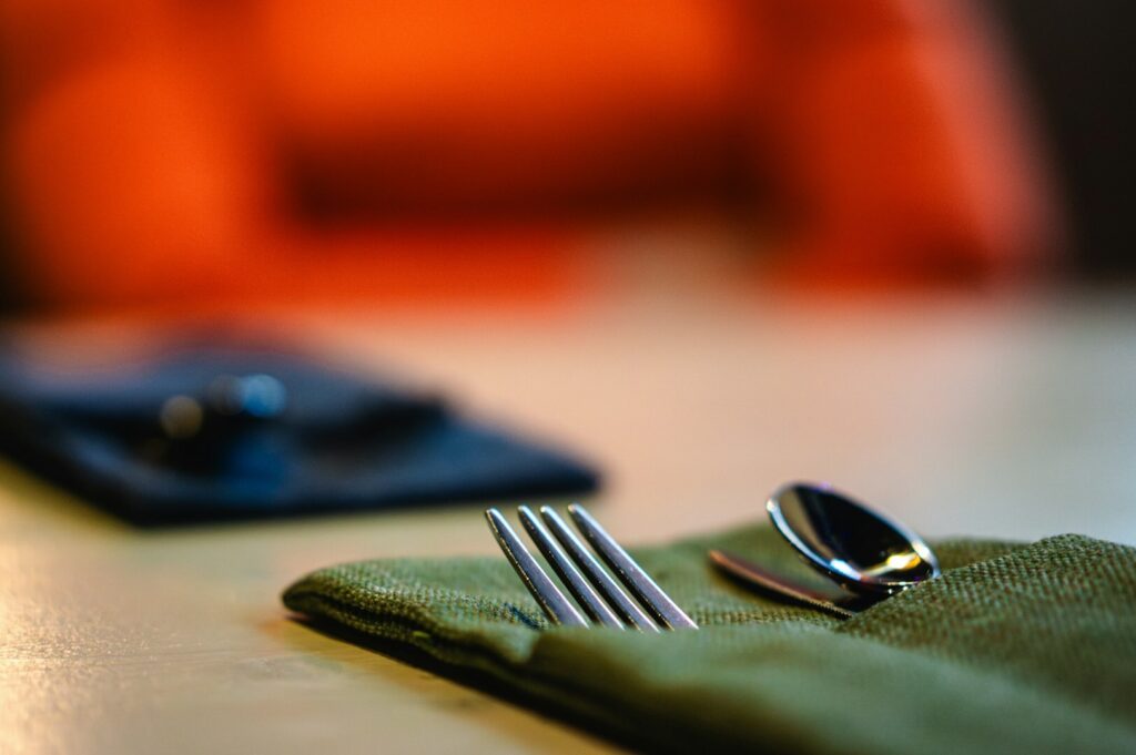 Close-up of a dining table setting with a fork and spoon on a green napkin in focus. A blurred orange chair provides a warm, cozy background.