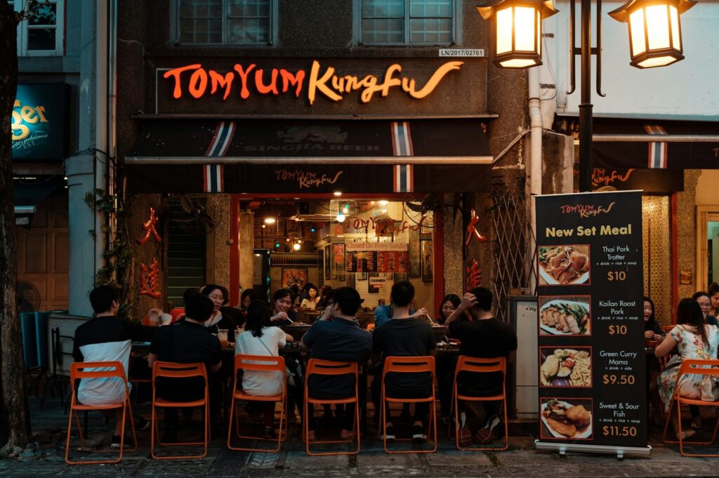 A group of people dine outdoors at "Tom Yum Kungfu" restaurant. Warm lighting and orange chairs create a cozy atmosphere. A menu board displays meal sets.