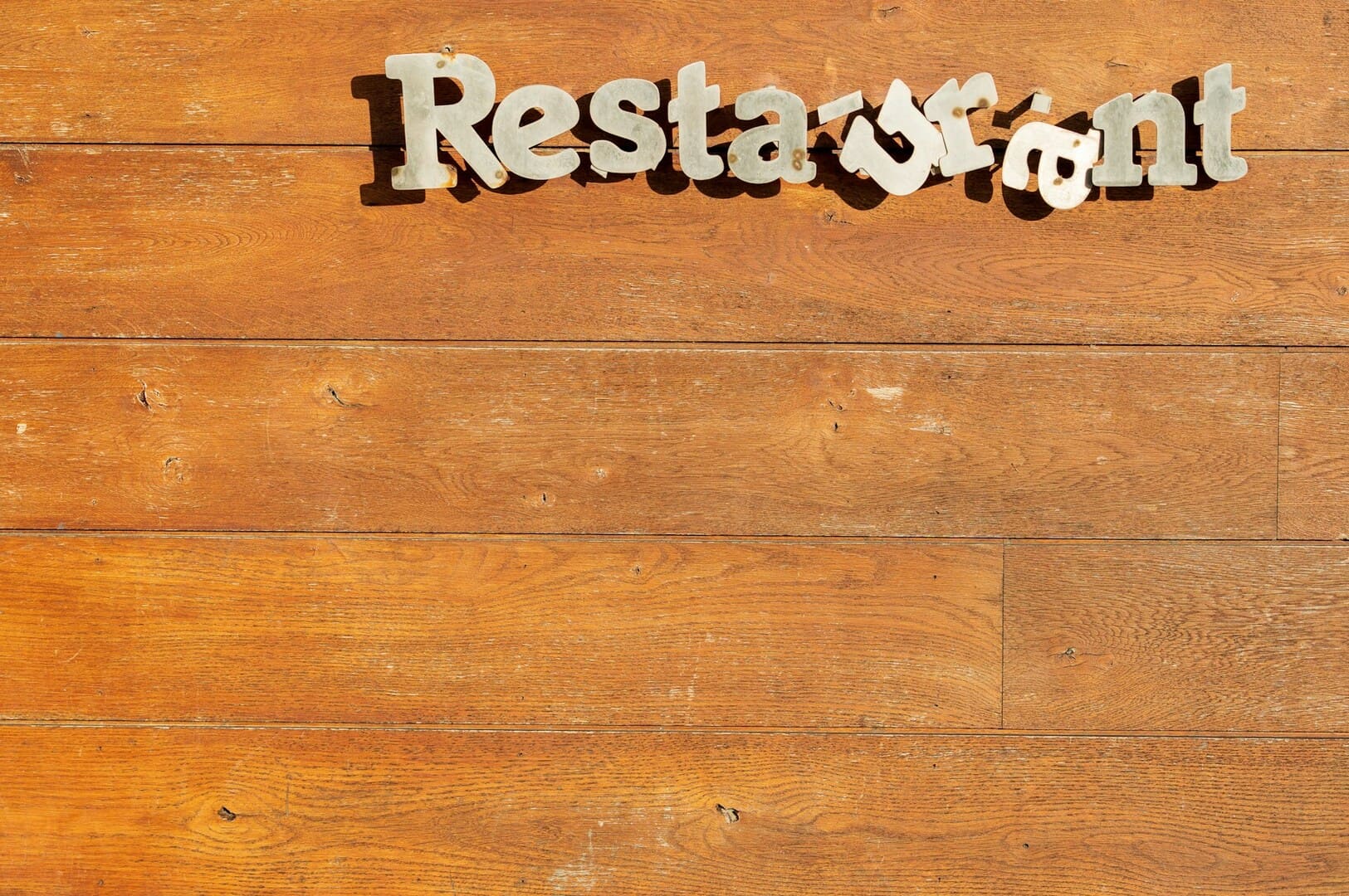 Metal letters spell "Restaurant" on a rustic wooden wall. The letters are slightly misaligned, adding a quirky charm to the scene