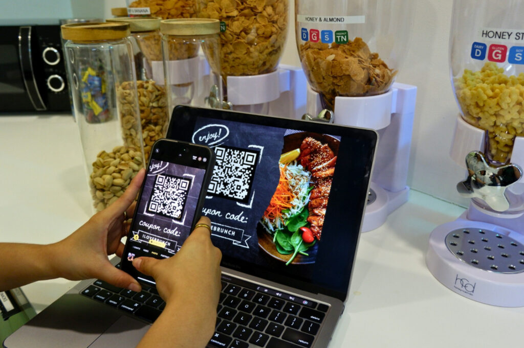 Hands hold a phone, scanning a QR code on a laptop displaying grilled meat and salad. Background features cereal dispensers, conveying a tech-savvy kitchen.