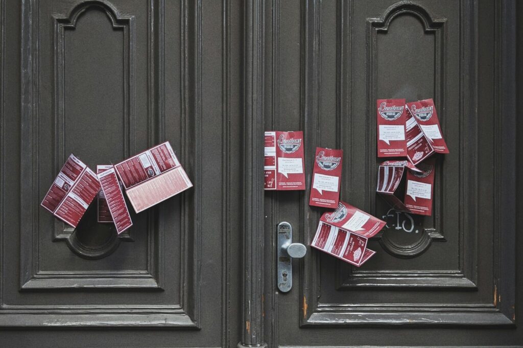 A dark wooden door with ornate panels, covered in numerous red flyers tucked into the handles and edges, suggesting clutter and neglect.