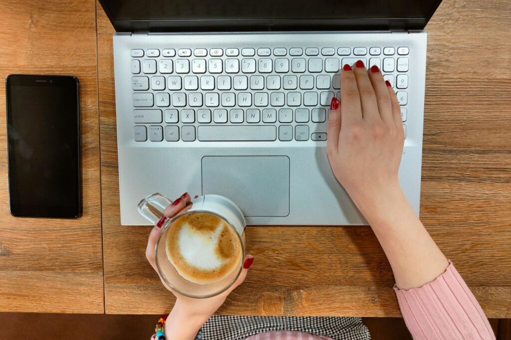 A person with red nails types on a laptop while holding a cappuccino. A smartphone rests nearby on a wooden table, creating a cozy work setting.