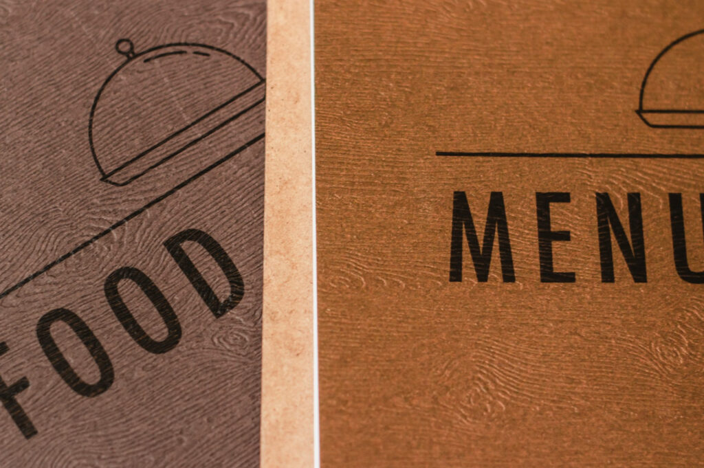 Close-up image of two brown menu covers. The left cover says "FOOD" with a cloche graphic, while the right says "MENU." Both have a textured wood pattern.