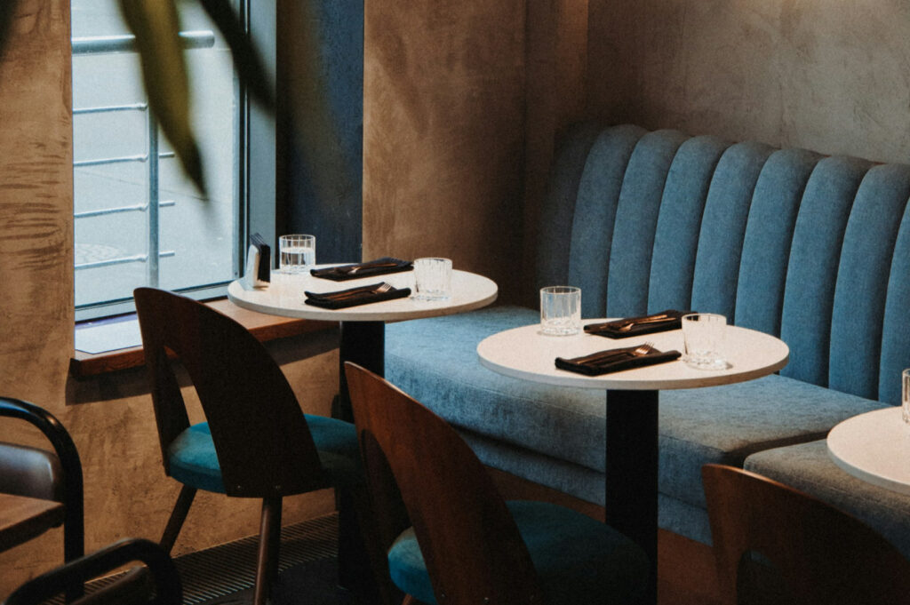 Cozy cafe setting with blue upholstered bench, wooden chairs, and two round tables set with glasses, black napkins, and cutlery by a large window.