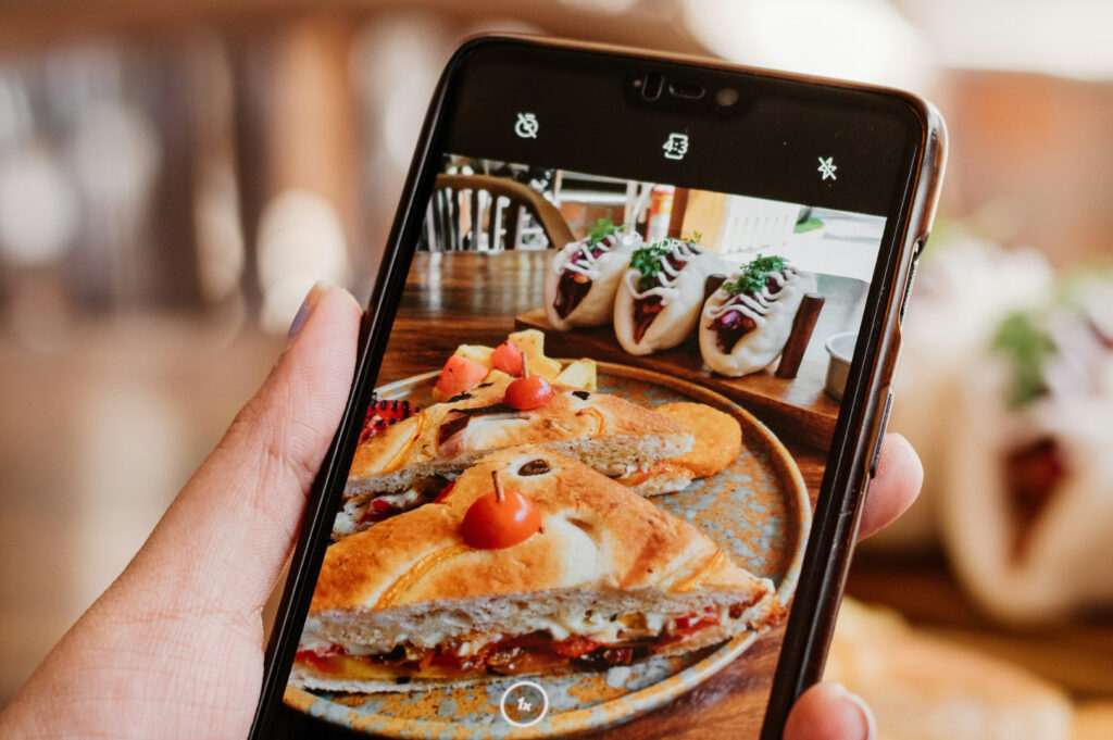 A hand holds a phone capturing a photo of a sandwich with cherry tomatoes and dips, with soft tacos visible in the background. The scene feels warm and inviting.