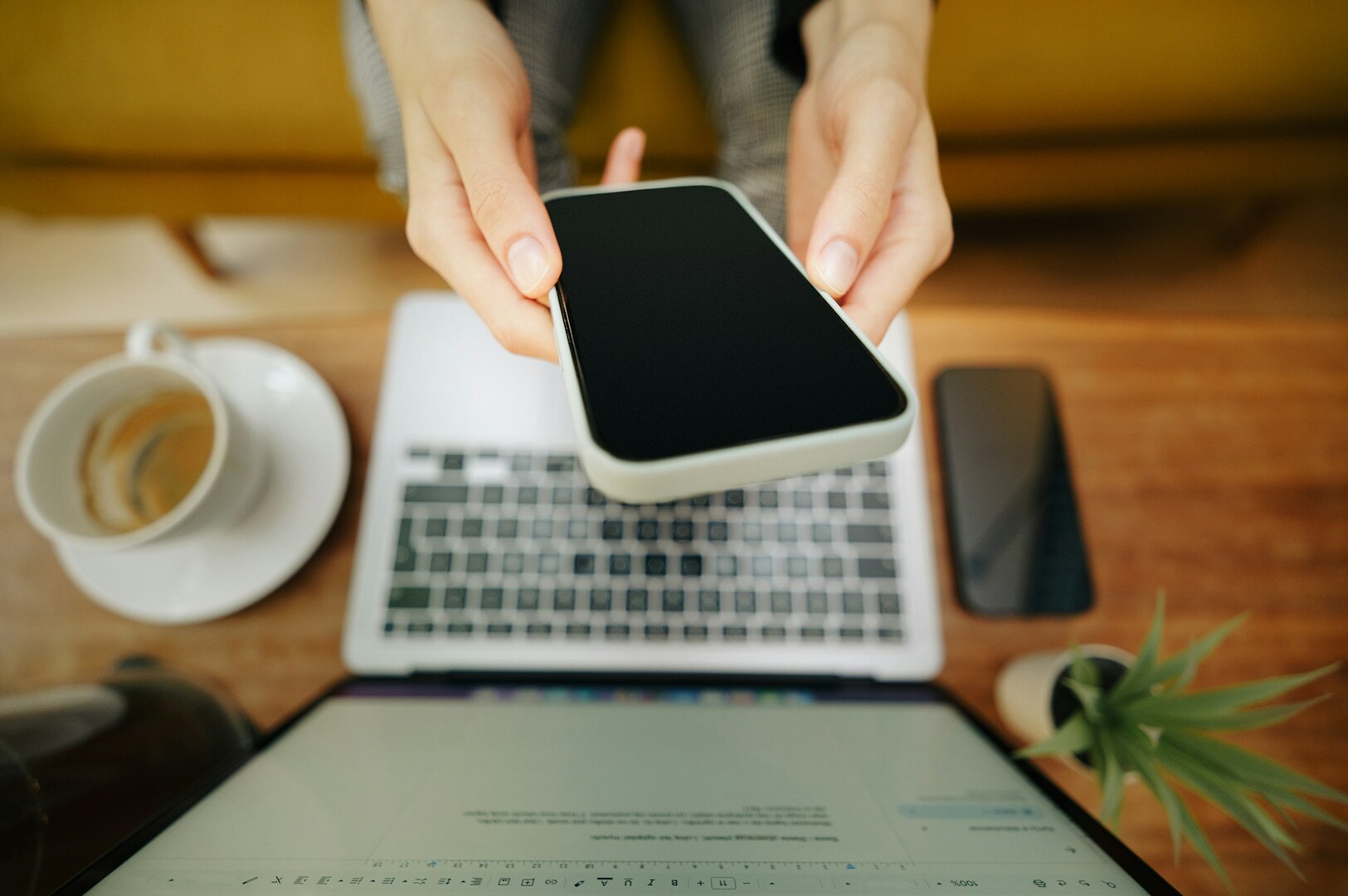 Hands hold a smartphone above an open laptop on a wooden desk. Nearby are a cup of coffee, another phone, and a small potted plant, conveying productivity.