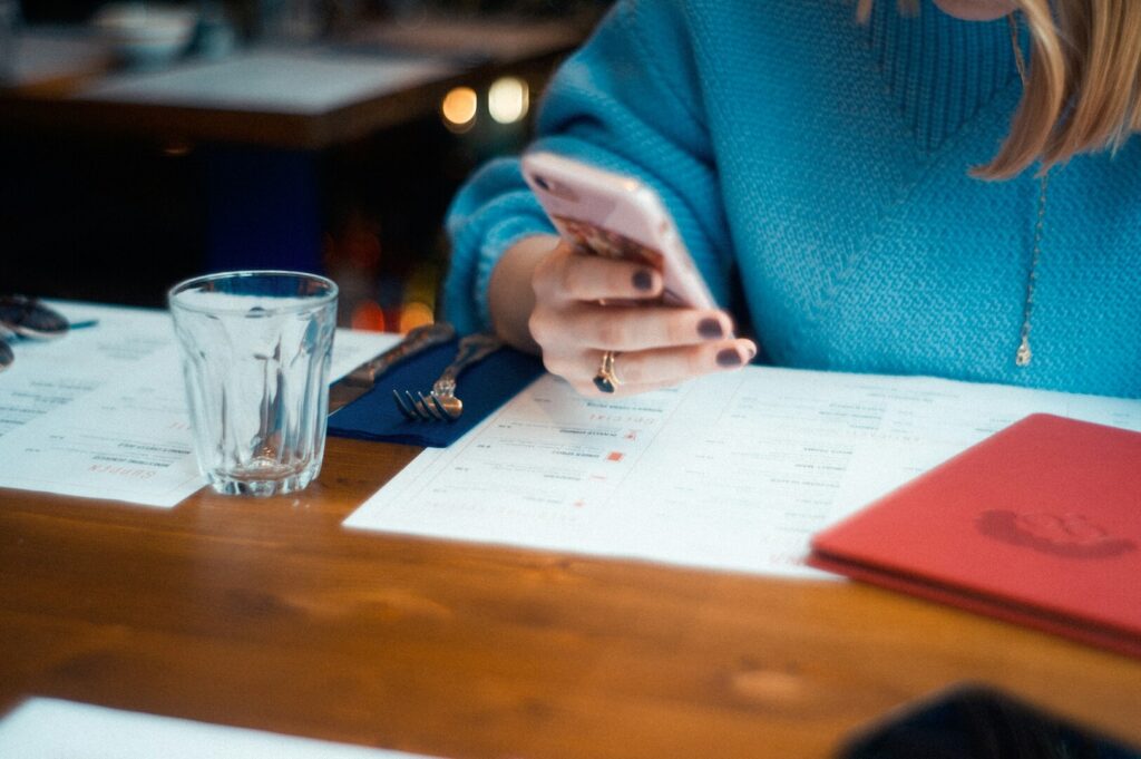 A person in a blue sweater is sitting at a wooden table, looking at a smartphone. A menu, glass, and utensils are nearby, creating a casual dining atmosphere.
