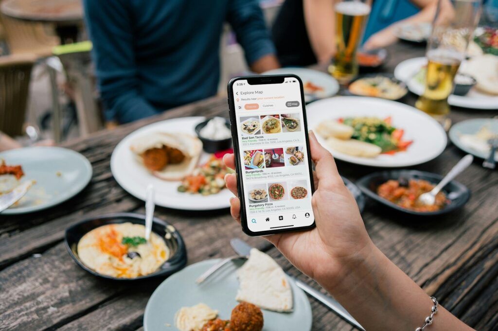 A person holds a smartphone displaying a restaurant app over a wooden table with plates of various Middle Eastern dishes and glasses of beer. Casual dining scene.