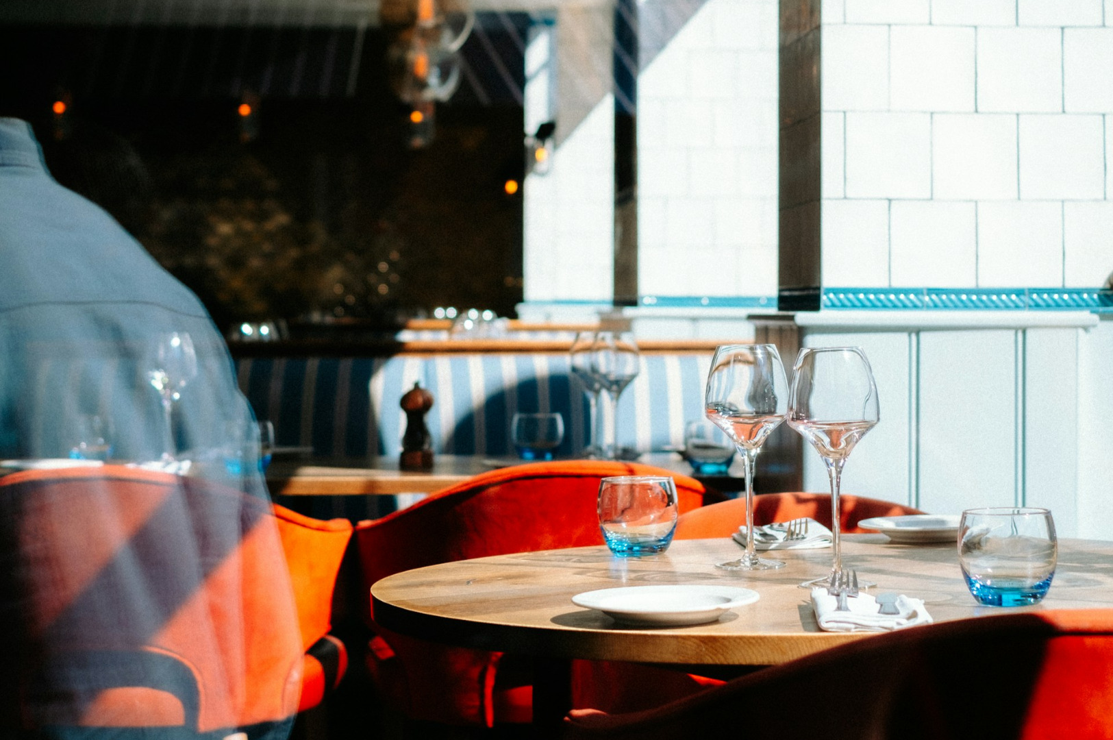 A cozy restaurant interior with a wooden table set with empty wine glasses, plates, and blue water glasses. Red cushioned chairs create a warm ambiance.