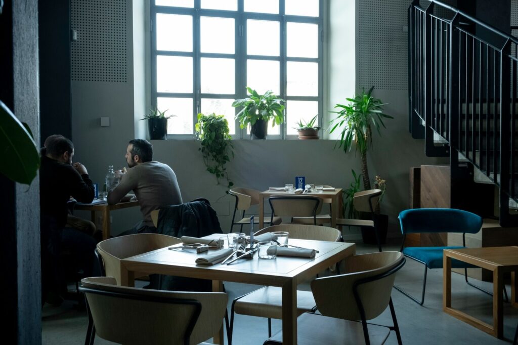 A cozy café with two people talking at a table by a large window. Indoor plants decorate the space; stairs are visible on the right. Quiet ambiance.