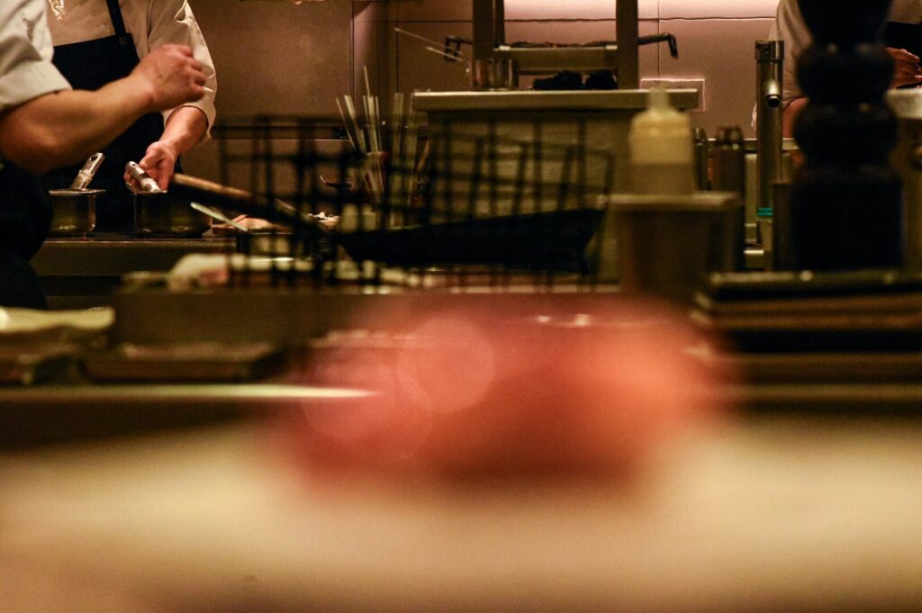 Chef in a dimly lit kitchen garnishing dishes, wearing a white shirt and black apron, surrounded by cooking utensils, conveying a focused atmosphere.