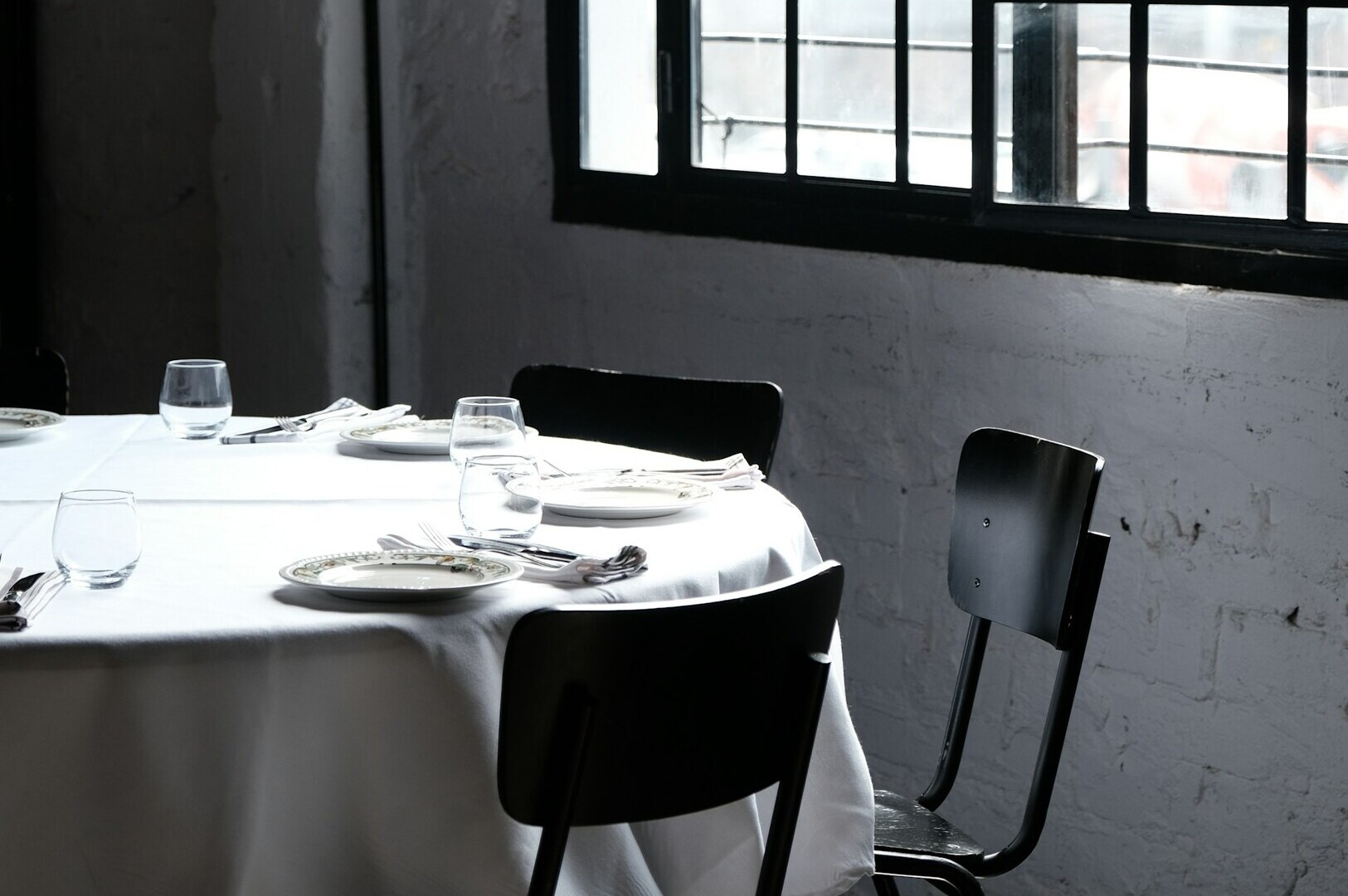 A round table set with white linens, plates, and glassware is surrounded by black chairs. Soft natural light enters through a grid-patterned window, creating a calm ambiance
