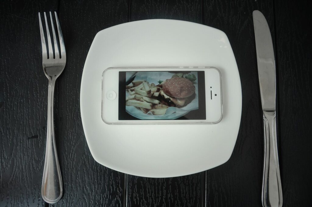 A smartphone displaying a burger and fries sits on a white plate, flanked by a fork and knife on a dark table, conveying modern dining and technology