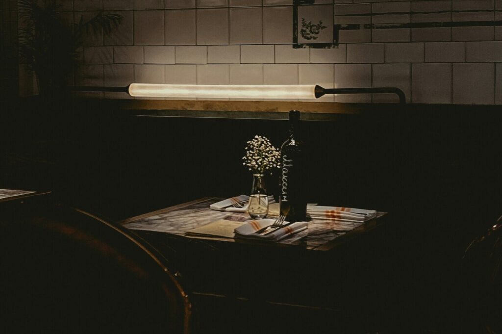 Cozy, dimly-lit restaurant table with a wine bottle, small white flowers in a vase, and neatly set silverware on napkins, evoking an intimate ambiance