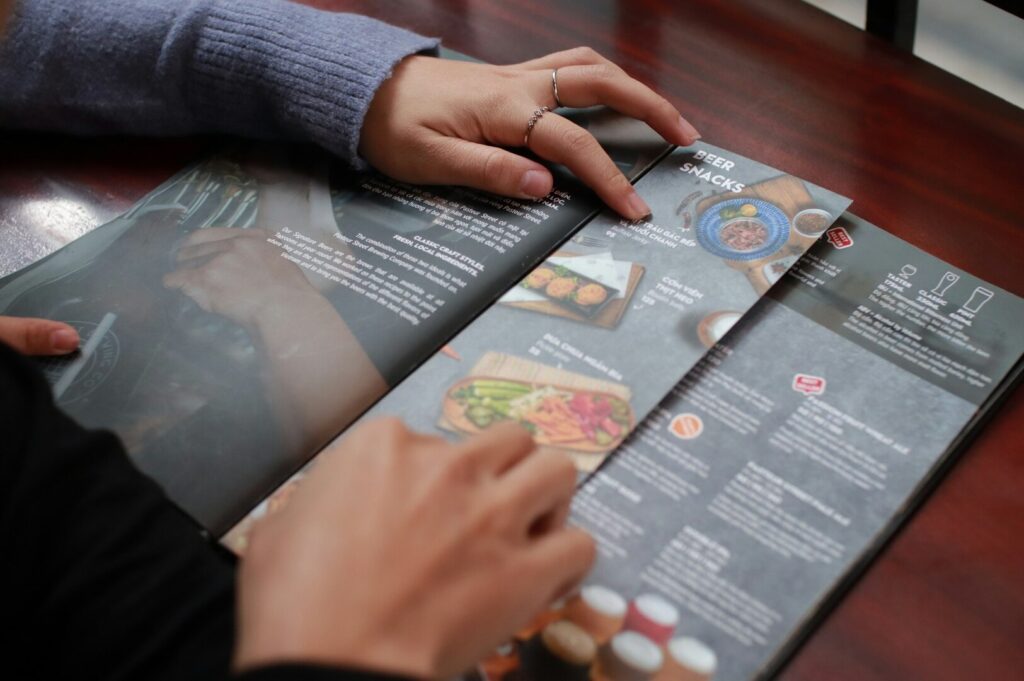 Two hands examining a restaurant menu on a wooden table. The menu displays images of food under the section “Beer Snacks,” suggesting a casual dining ambiance.
