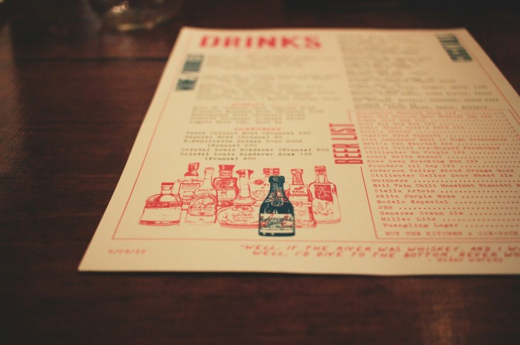 A vintage-style drink menu on a wooden table features illustrations of bottles with red and teal accents. The mood is rustic and inviting.