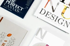 Four design booklets arranged on a white surface. Top left: dark blue cover with "The Perfect Canvas." Top right: "Design" with utensil graphics. Bottom left: floral pattern. Bottom right: "Neenah" on a sleek white cover