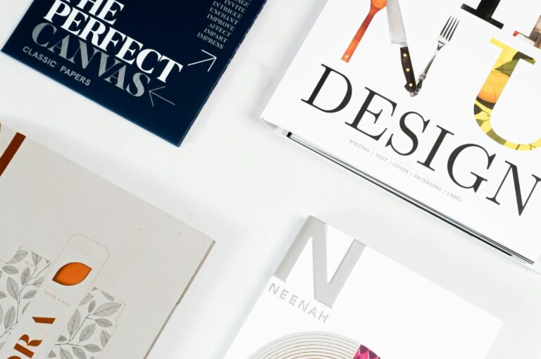 Four design booklets arranged on a white surface. Top left: dark blue cover with "The Perfect Canvas." Top right: "Design" with utensil graphics. Bottom left: floral pattern. Bottom right: "Neenah" on a sleek white cover