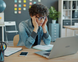 Woman sitting in front of a laptop with a stressed expression, surrounded by work materials, appearing overwhelmed by tasks.