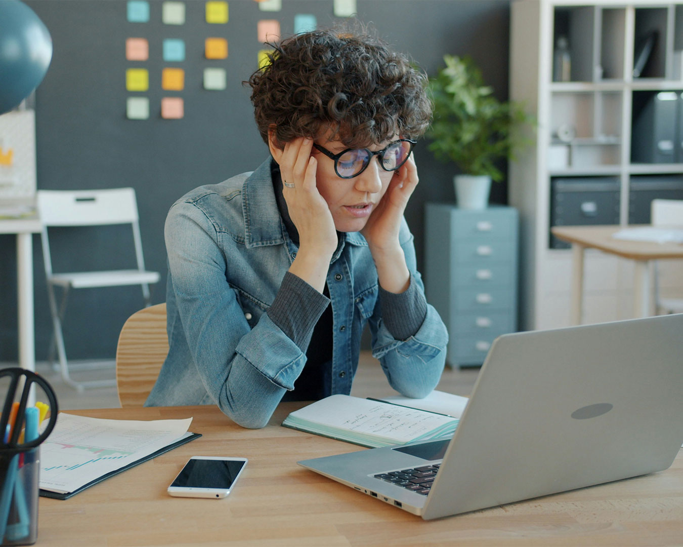 Woman sitting in front of a laptop with a stressed expression, surrounded by work materials, appearing overwhelmed by tasks.
