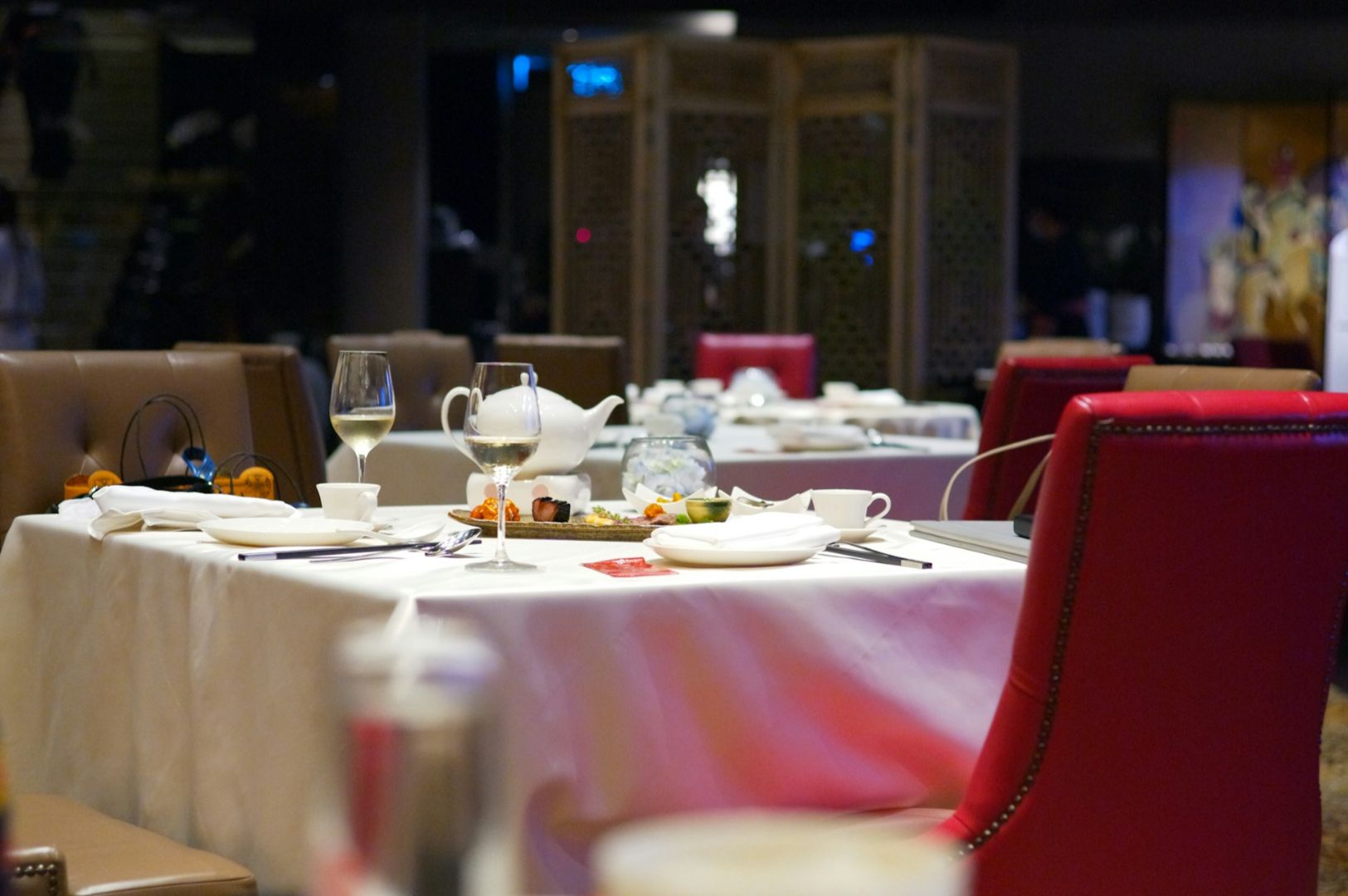 Elegant dining table set with white tablecloth, glass of wine, tea set, and a plate of appetizers. Red and brown chairs add a warm, inviting tone.