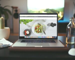Laptop on a desk showing a food blog. Nearby are a cactus, bottled water, a mouse, and a newspaper. Background features a blurred screen, creating a cozy ambiance.