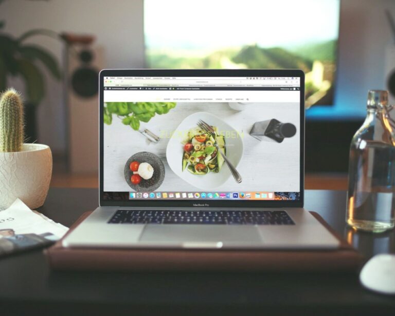 Laptop on a desk showing a food blog. Nearby are a cactus, bottled water, a mouse, and a newspaper. Background features a blurred screen, creating a cozy ambiance.
