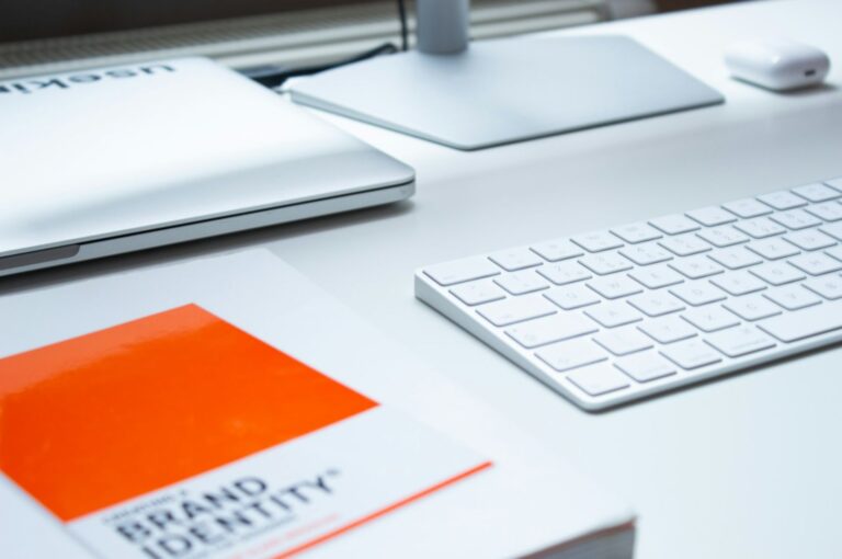 "A clean, organized workspace with a white keyboard, closed laptop, and a bright orange "Brand Identity" book on the desk, conveying a modern, professional tone."
