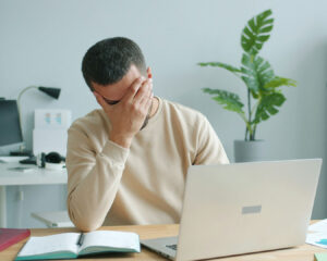 A man sitting at his desk with a tired expression, surrounded by a laptop and papers, showing signs of work stress and pressure.
