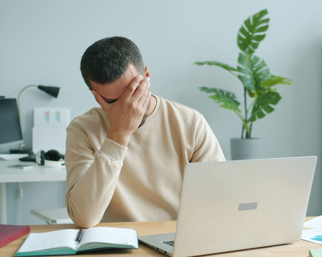 A man sitting at his desk with a tired expression, surrounded by a laptop and papers, showing signs of work stress and pressure.