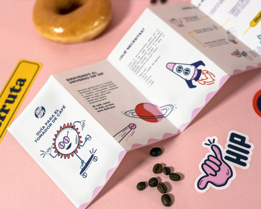 A colorful brochure on a pink surface, featuring playful illustrations of a rocket and a sun with glasses. Surrounded by coffee beans, a donut, and stickers.