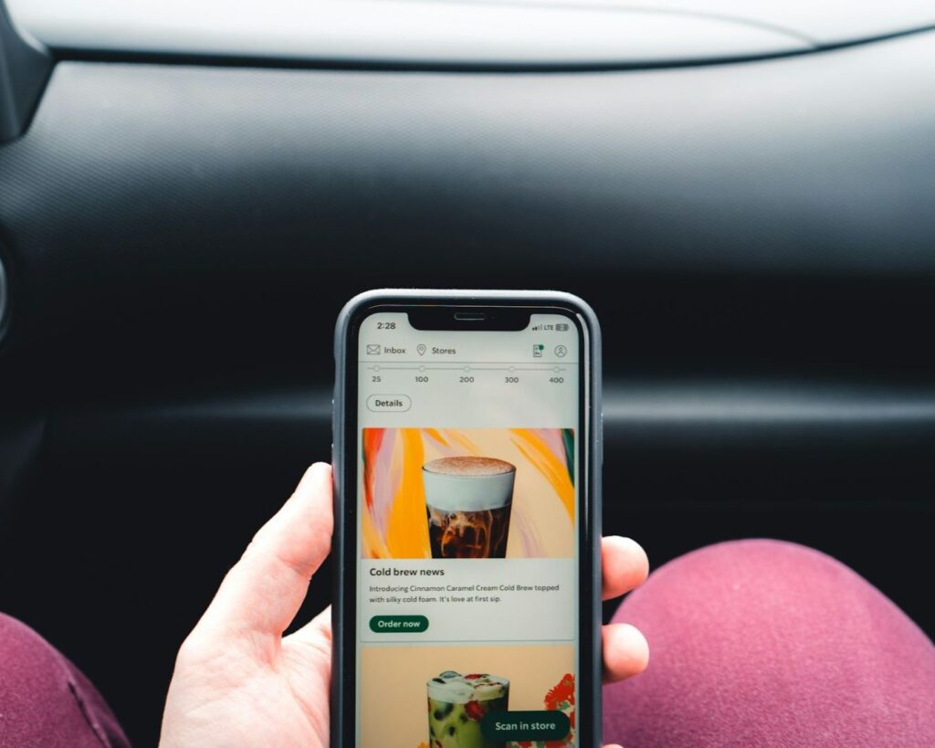 Hand holding a smartphone in a car, displaying an app with images of iced coffee and vibrant colors. Dashboard visible in the background. Casual tone.