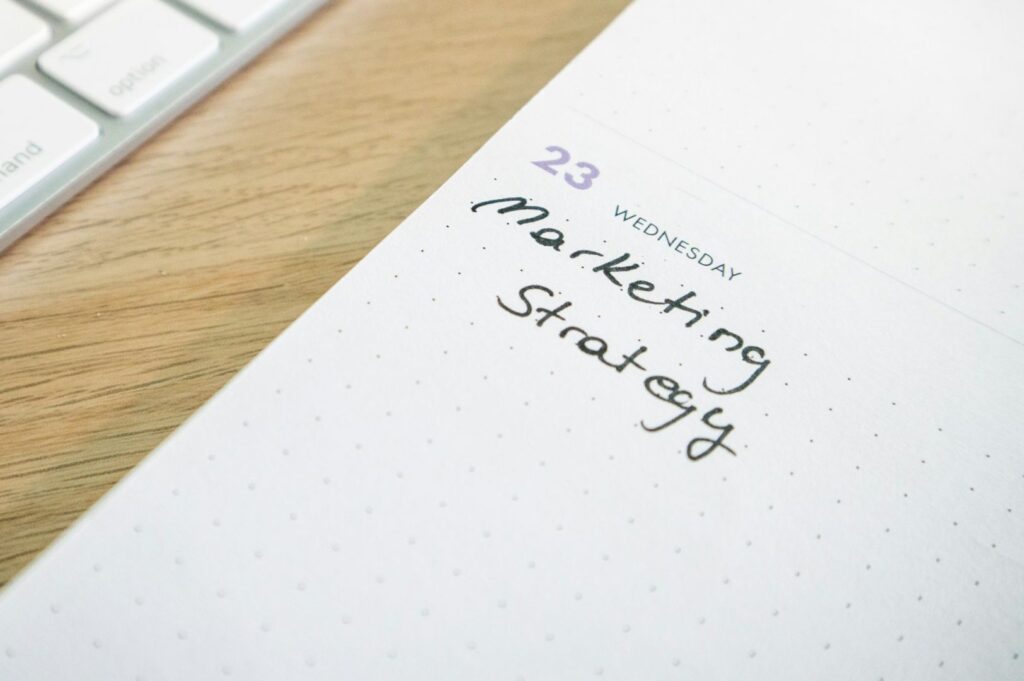 Close-up of an open planner on a wooden desk, showing "Wednesday 23" with "Marketing Strategy" handwritten in black ink. A computer keyboard is partially visible.
