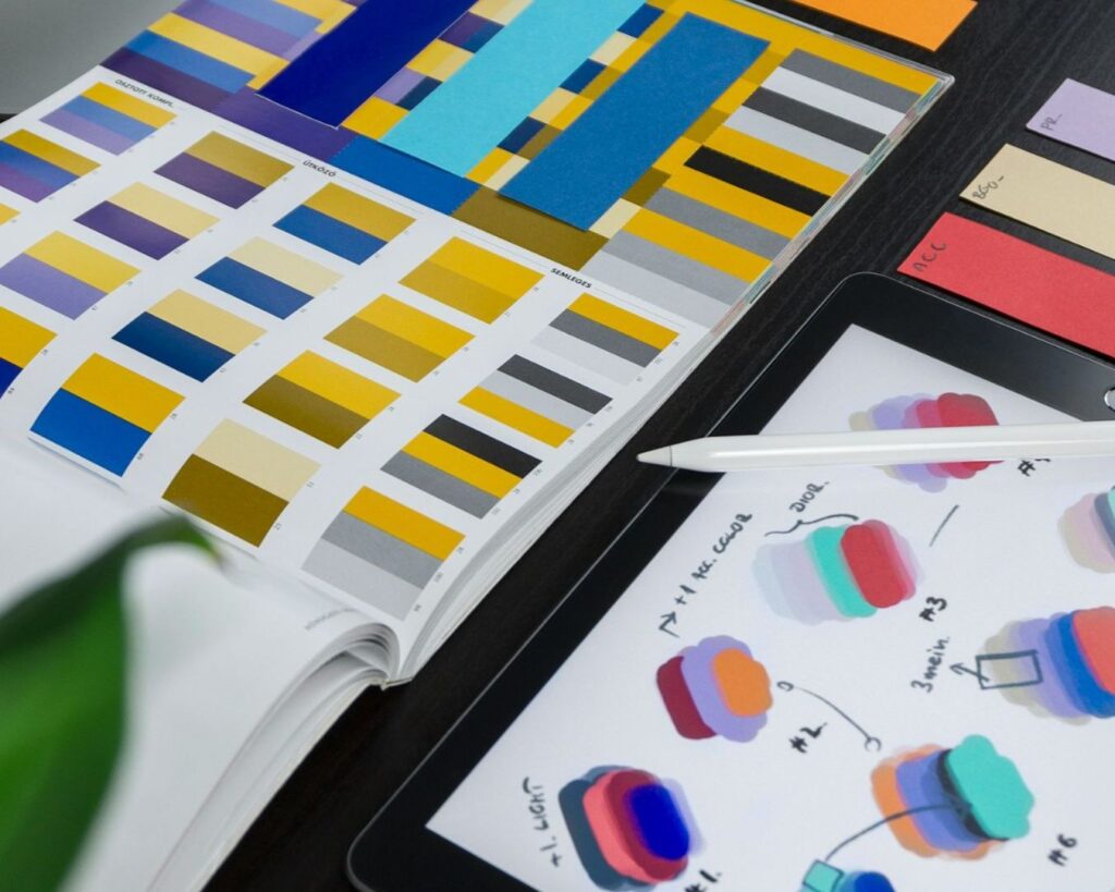 Open color swatch book with vibrant yellow, blue, and gray samples. Nearby, a digital tablet displays colorful cloud-like graphics and notes.