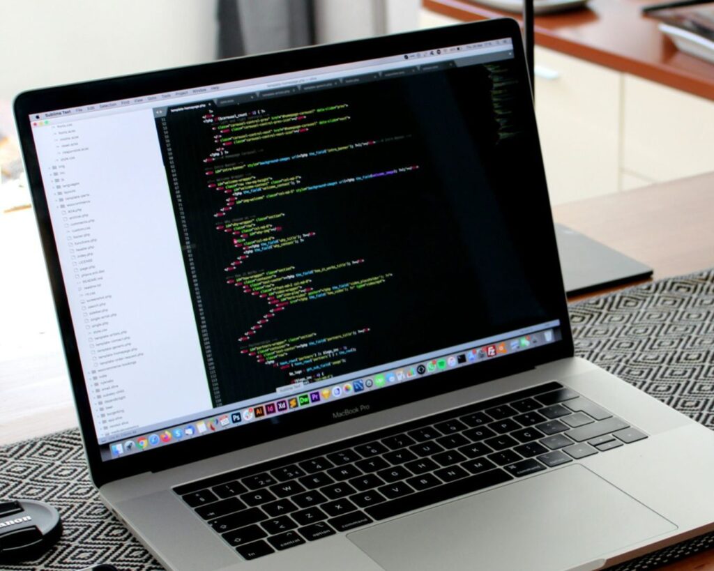 A laptop displaying colorful code on a screen sits on a patterned table. Nearby are a camera lens cap, a mouse, and a modern, organized desk setup
