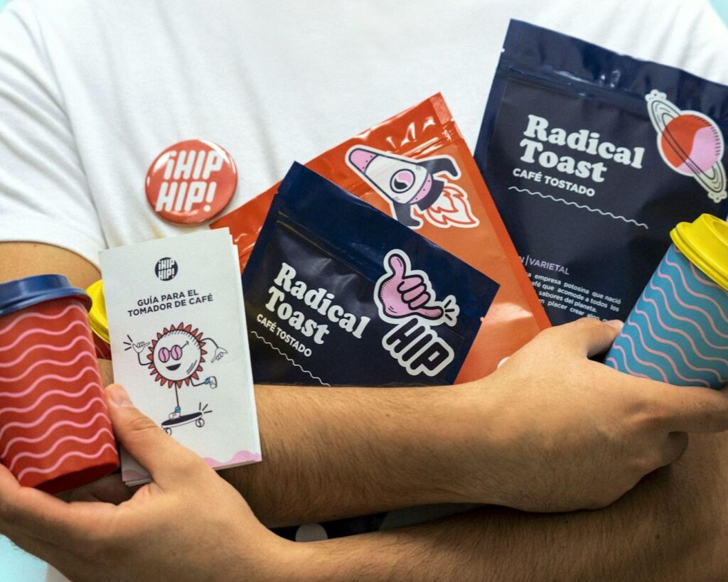 A person holds colorful coffee bags and cups, wearing a "HIP HIP!" button. The vibrant packaging and playful illustrations create a cheerful and energetic vibe.