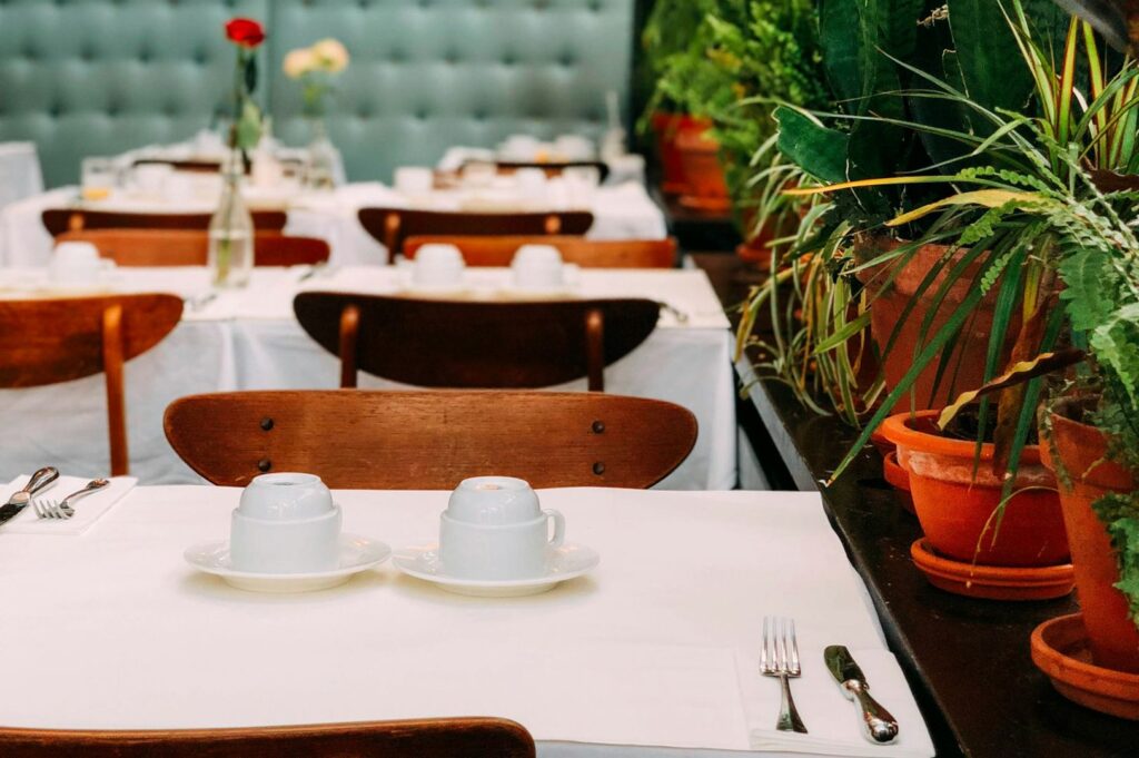 Elegant restaurant setting with white tablecloths, arranged china, and wooden chairs. Potted plants and flowers add greenery and warmth to the scene.