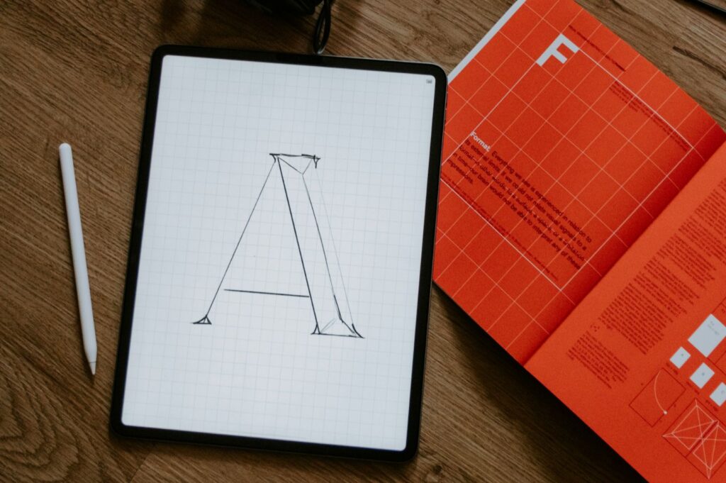 A digital tablet on a wood surface displays a sketched letter "A" with grid lines. Nearby, an open orange typography book and a white stylus are visible.