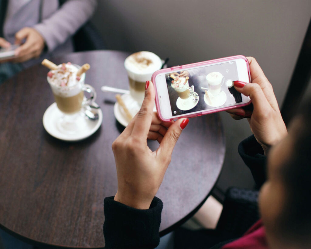 A woman holding her phone above a cup of coffee, capturing a photo of the drink at a café table with warm lighting and a relaxed atmosphere.