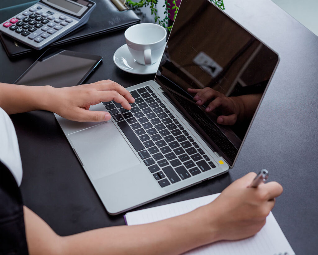 A hand writing notes while the other types on a laptop keyboard, showing multitasking and active work in progress.