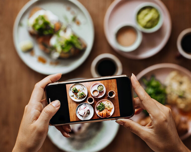 A smartphone held above a plated dish, capturing a close-up food photo under warm restaurant lighting, highlighting the textures and colours of the meal.