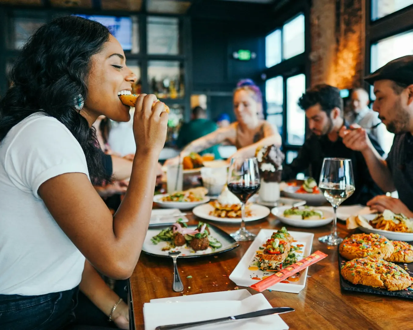 A group of friends enjoys a lively meal at a restaurant. A woman in the foreground bites into food, surrounded by colorful dishes and drinks, conveying joy.