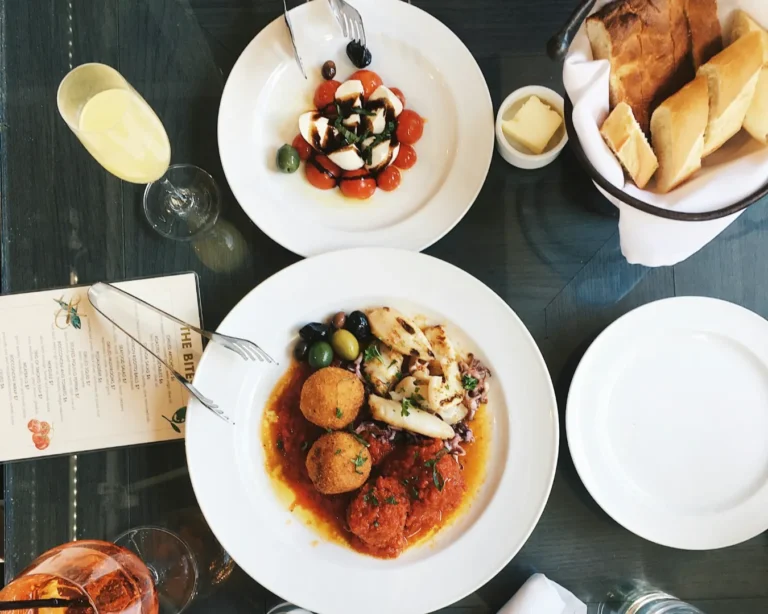 A table setting features a Caprese salad with tomatoes and mozzarella, a main dish with arancini and olives, bread with butter, a menu, and a glass of orange juice.