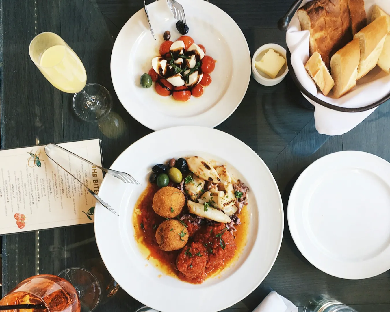A table setting features a Caprese salad with tomatoes and mozzarella, a main dish with arancini and olives, bread with butter, a menu, and a glass of orange juice.