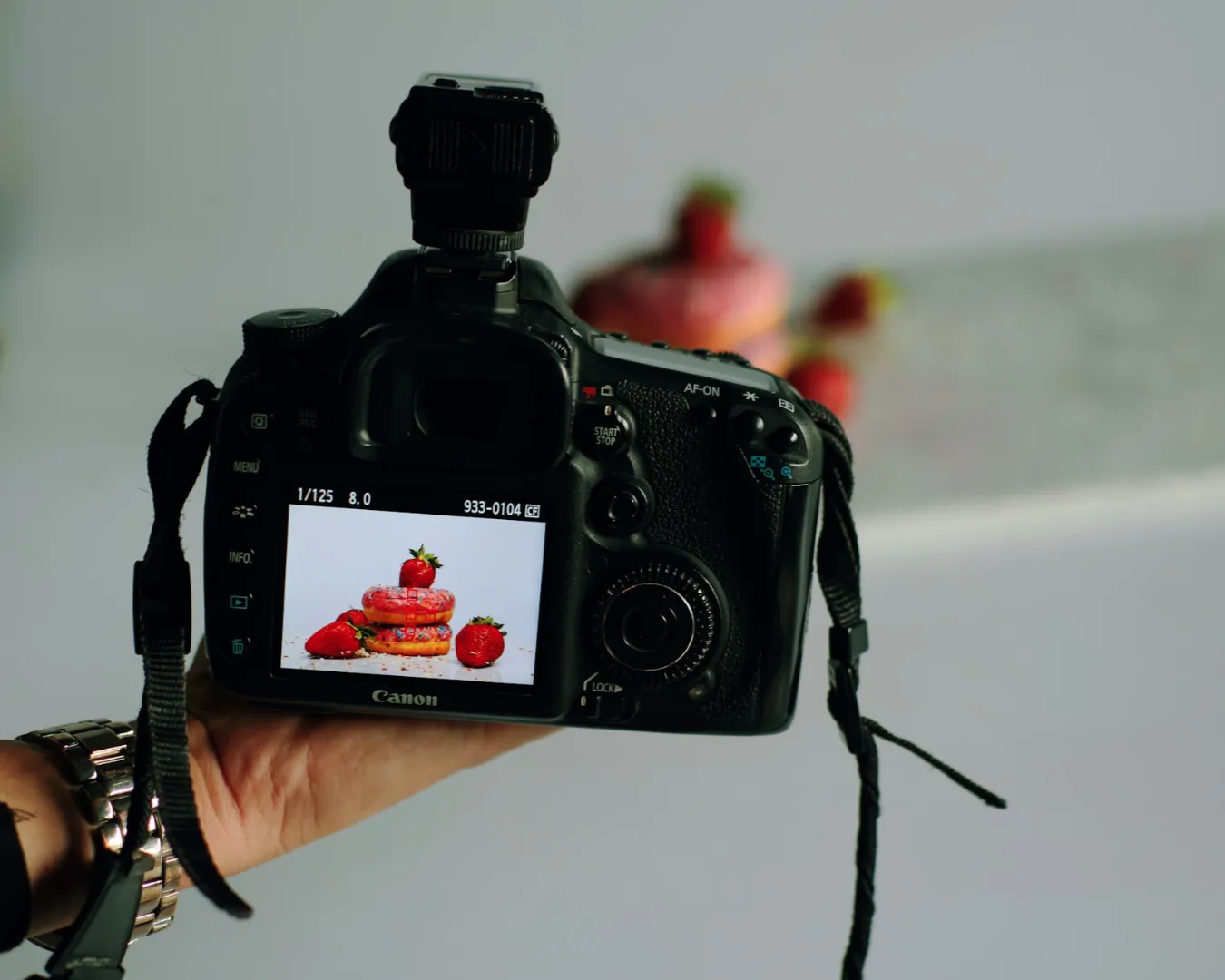A DSLR camera screen displays a stack of pink macarons with strawberries on top. The setup is in a studio, conveying a professional, creative tone.