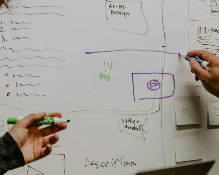 Two people collaborate at a whiteboard covered with sketches and notes in various colors. Their hands gesture, indicating brainstorming and planning.