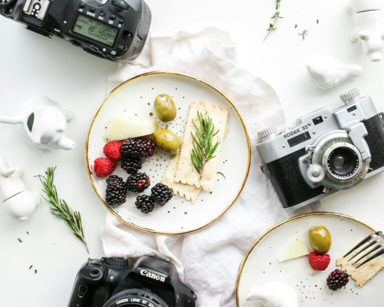 Aerial view of a minimal table scene with vintage cameras, two plates of olives, crackers, cheese, berries, rosemary, and white ceramic figurines. Bright and artistic.