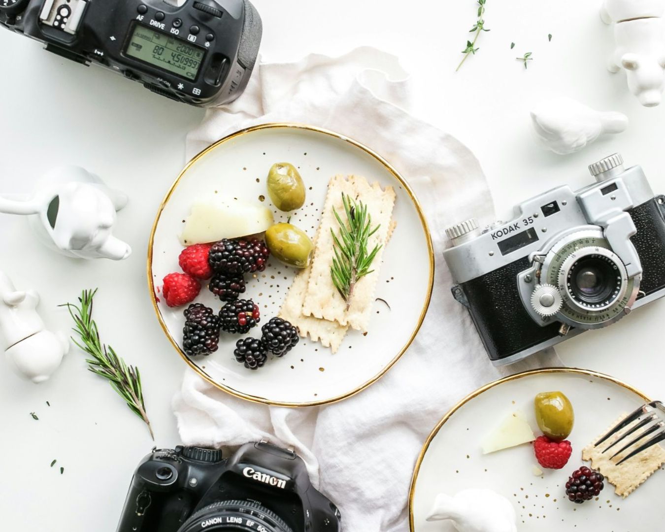 Aerial view of a minimal table scene with vintage cameras, two plates of olives, crackers, cheese, berries, rosemary, and white ceramic figurines. Bright and artistic.