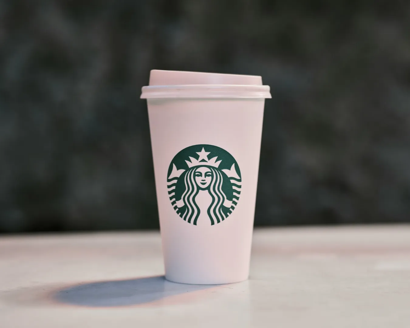 A white Starbucks cup with a green logo sits on a table against a blurred dark background, conveying a minimalist and calm atmosphere.